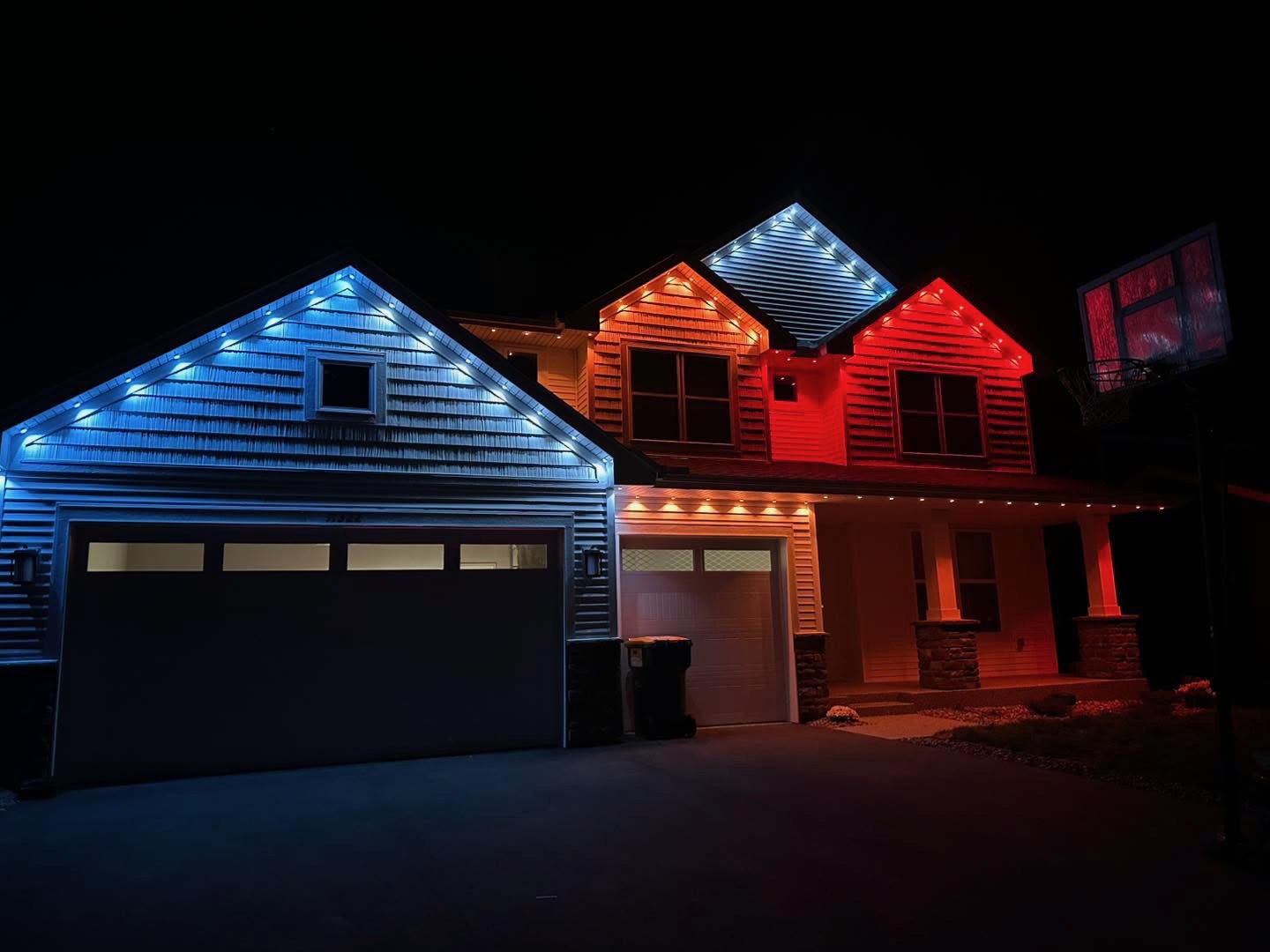 Two-story home at night with a multi-color holiday theme — blue, amber, white and red across the gables and porch